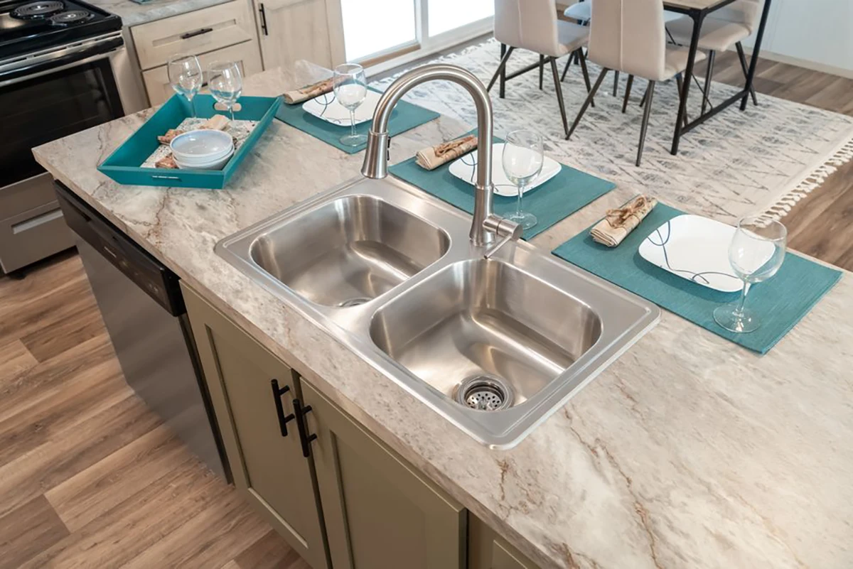 A double-basin stainless steel sink with a curved faucet is set in a marble countertop. Place settings with teal placemats and wine glasses are arranged nearby.