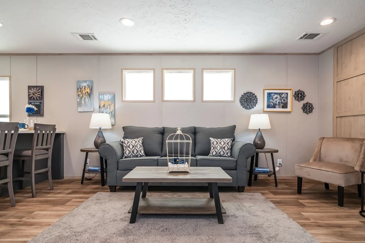 A living room with a gray sofa, patterned pillows, a coffee table, a beige armchair, wall art, lamps, and three high windows above the sofa.