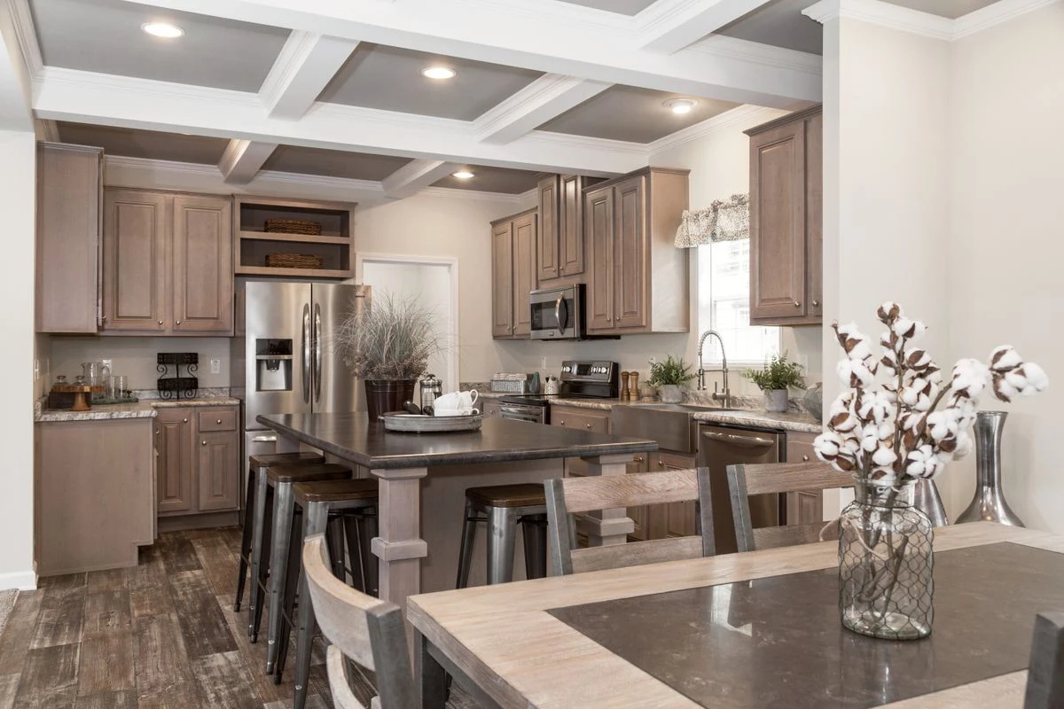 Modern kitchen with wooden cabinets, stainless steel appliances, a central island with seating, and a dining table in the foreground. Decorative plants and cotton stems are visible.