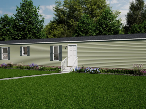 A single-story, light green manufactured home with white trim, small steps leading to the front door, and a well-kept lawn bordered by shrubs and flowers, set against a backdrop of trees.