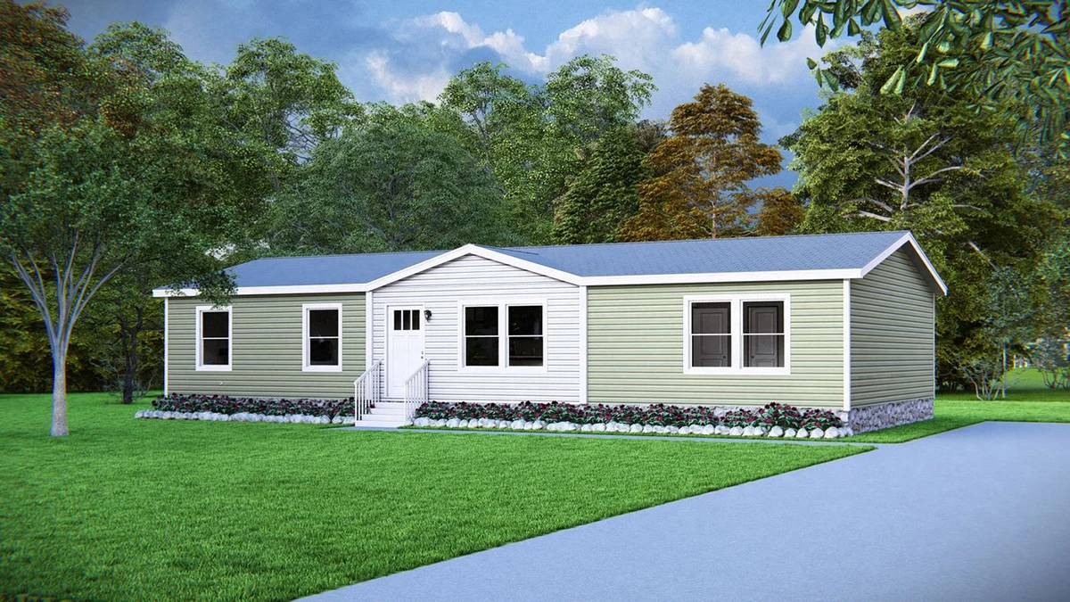 A single-story manufactured home with a gabled roof, light green siding, white trim, front steps, and a small flower bed, set on a grassy lawn with trees in the background.