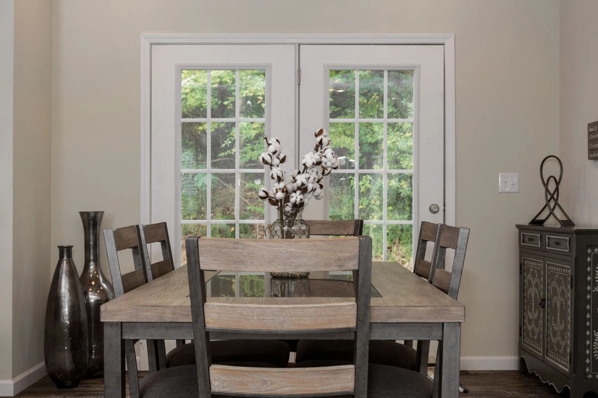 Dining room with a wooden table and six chairs, a vase with cotton stems as centerpiece, and French doors opening to a green outdoor area.
