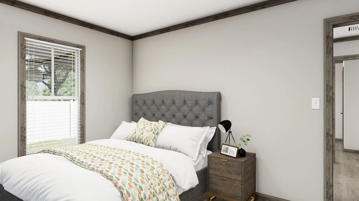 A bedroom with a gray upholstered bed, patterned bedding, a wooden nightstand holding a lamp, a photo frame, and a plant, next to a window with blinds.