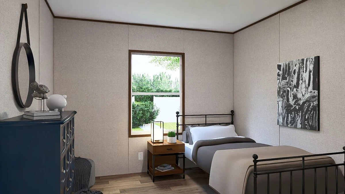 A neatly made single bed sits beside a window in a simple bedroom with neutral walls, wooden floors, a blue dresser, and a nightstand with a lamp and books.
