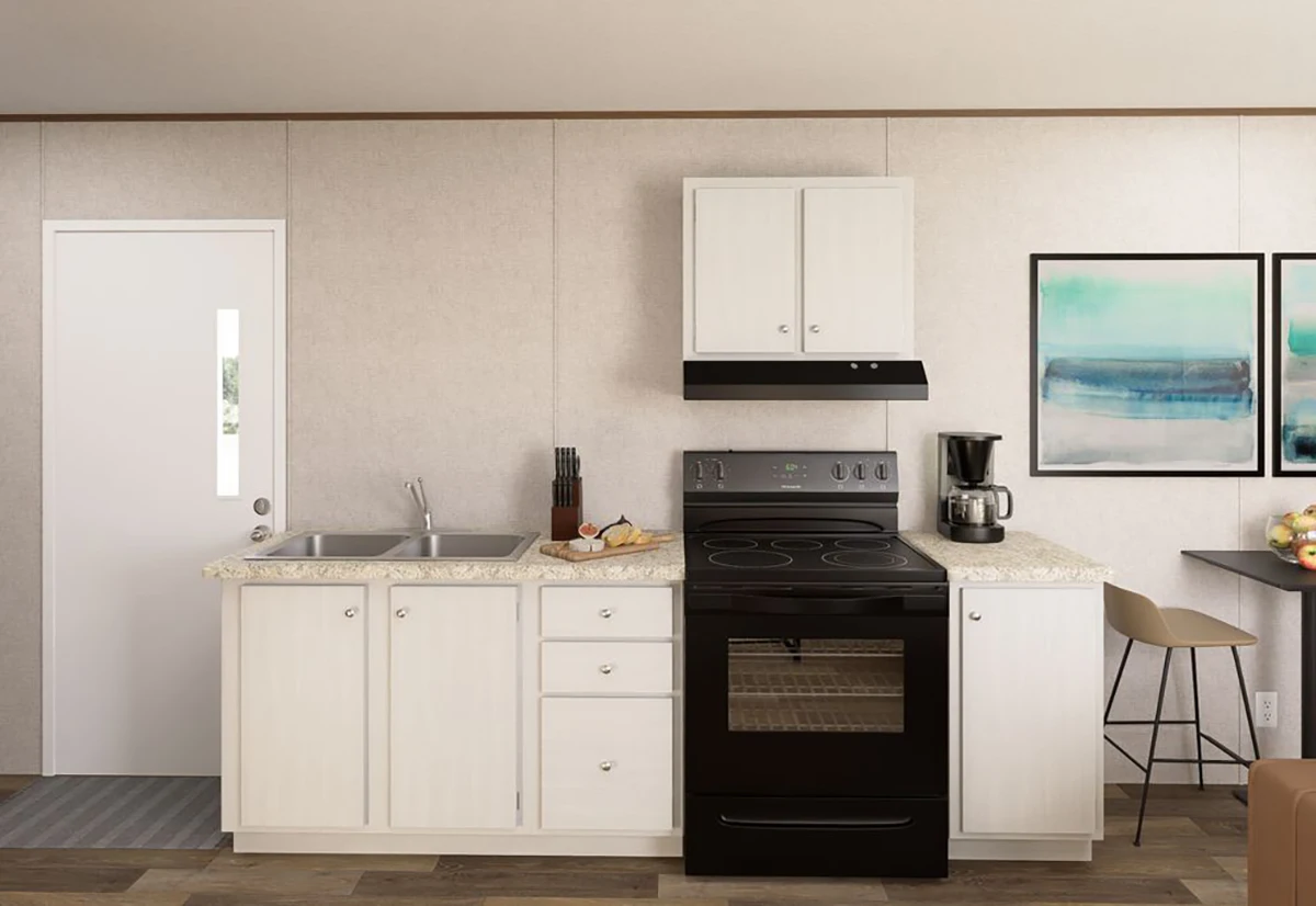Compact kitchen with white cabinets, black stove, coffee maker, sink, countertop items, wall art, and a door to the left.