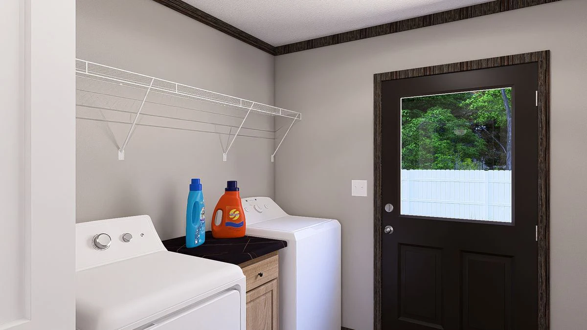 1fb3876e-6b57-42b2-b84e-6b3b1cebb1cb A laundry room with a washing machine, dryer, bottles of detergent on a counter, a wire shelf above, and a door with a window showing a white fence and greenery outside.