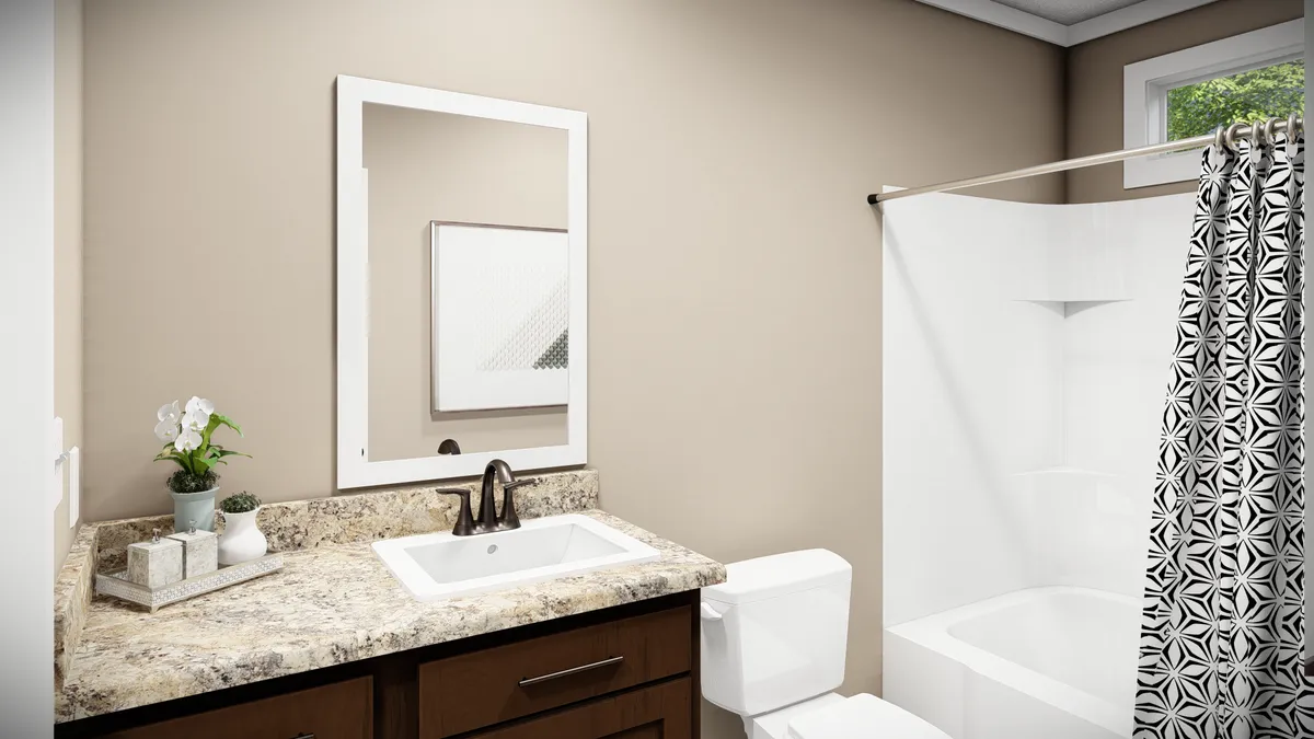 Bathroom with a beige wall, white-framed mirror, granite countertop sink, toilet, potted plant, and a white bathtub with a patterned shower curtain.