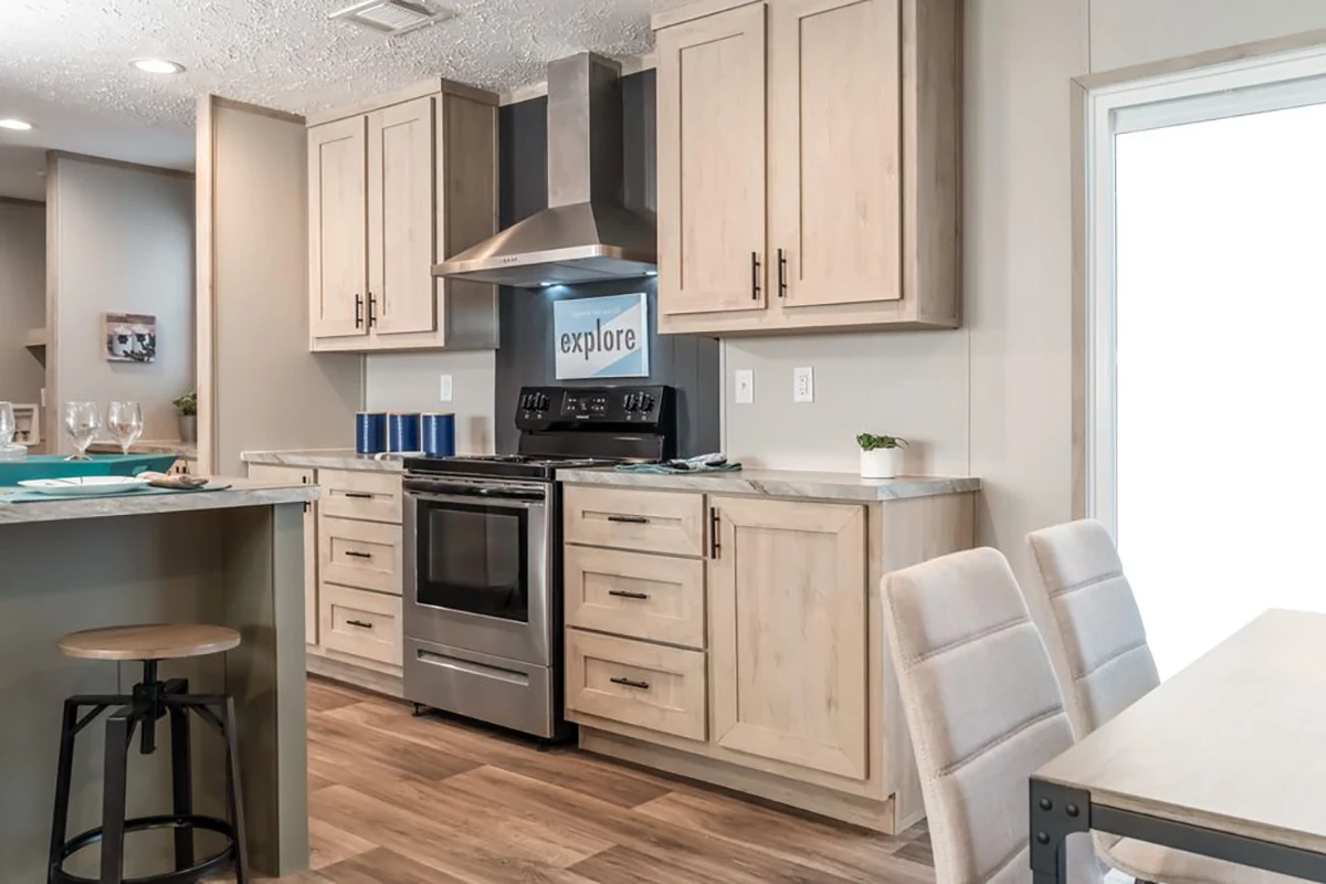 Modern kitchen with light wood cabinets, stainless steel appliances, range hood, island with a stool, and a dining table with upholstered chairs.