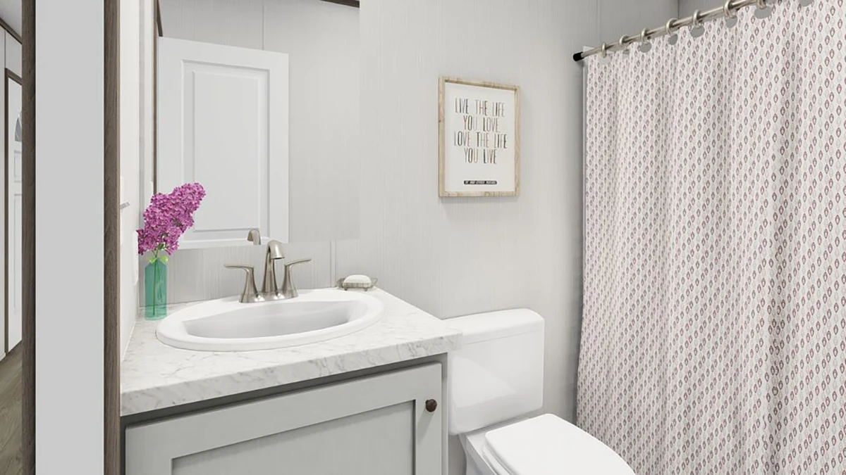 Modern bathroom with a white sink, marble countertop, and silver faucet. A vase with purple flowers sits by the mirror. There is a white toilet and a shower with a patterned curtain.