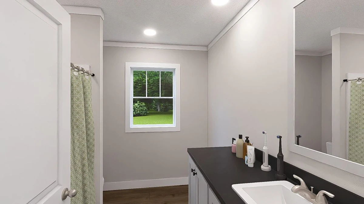 A bathroom with a sink, countertop items, a large mirror, a window showing greenery outside, and a shower with a green-patterned curtain.