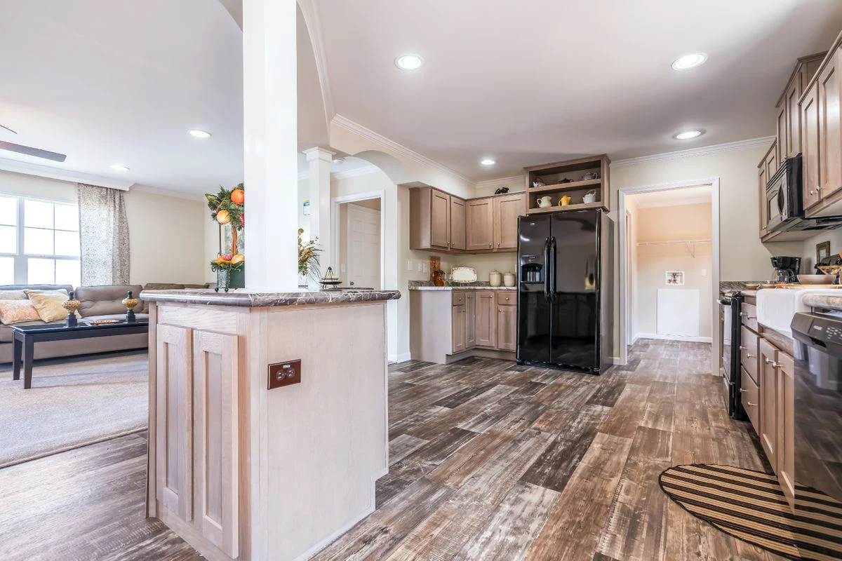 Open kitchen with wood cabinets, black appliances, and brown patterned flooring; adjacent living area with a sofa and large window visible in the background.