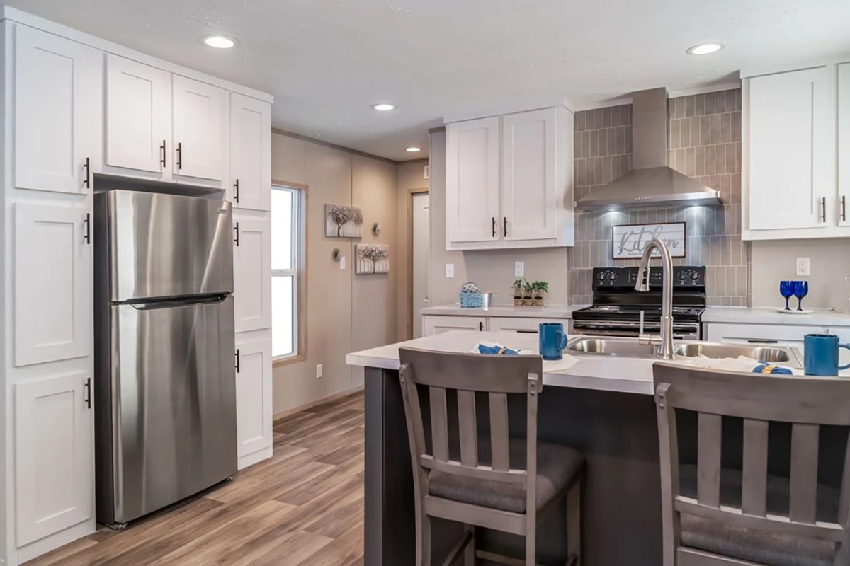 Modern kitchen with white cabinets, stainless steel appliances, an island with a sink, and two gray chairs. Recessed lighting and wooden flooring complete the space.