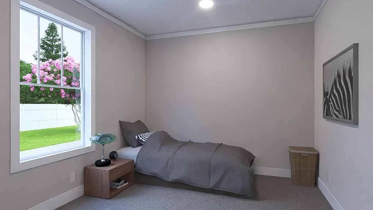 A small, simply furnished bedroom with a single bed, bedside table, lamp, framed wall art, laundry basket, and a window overlooking a yard with a flowering tree.