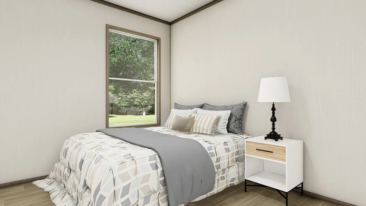 A neatly made bed with patterned bedding, several pillows, and a gray throw sits beside a white nightstand with a lamp in a simple bedroom with natural light from a window.