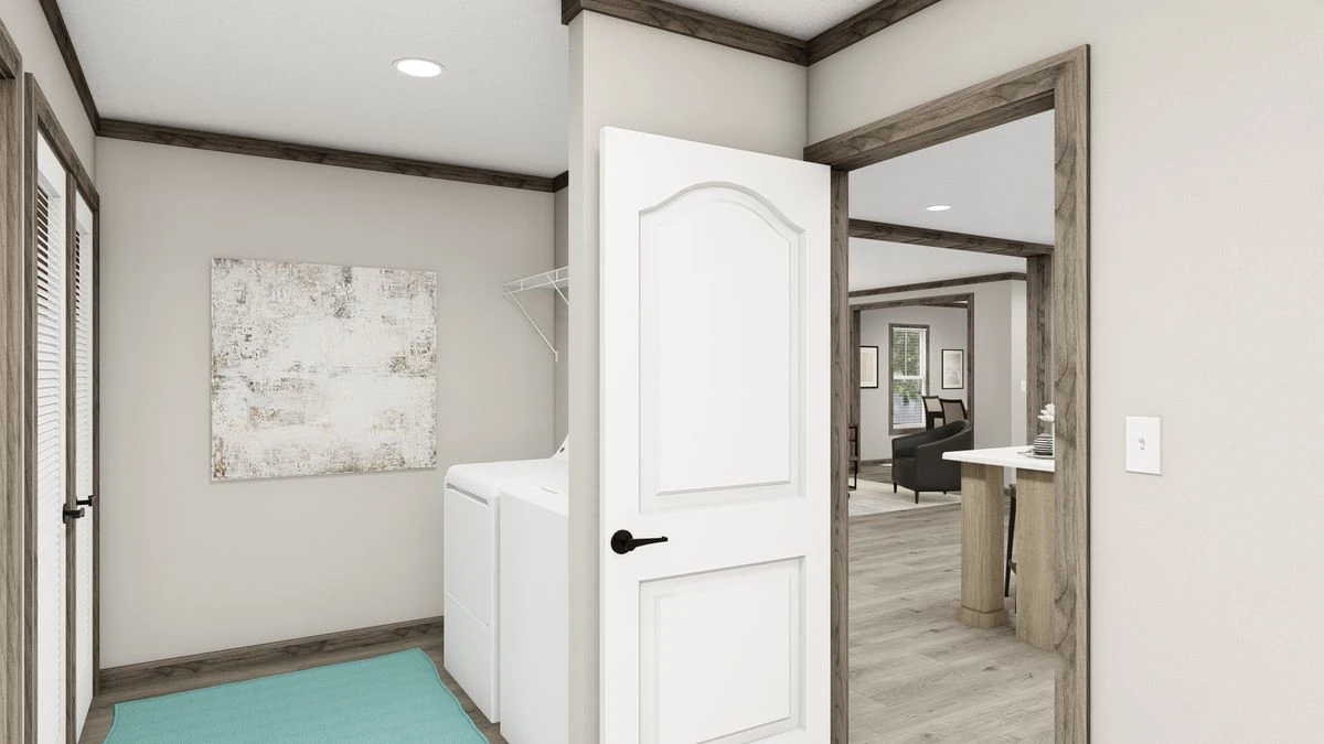 A laundry room with a washing machine, open door, wall art, and a turquoise mat, leading to a hallway with light wood flooring.