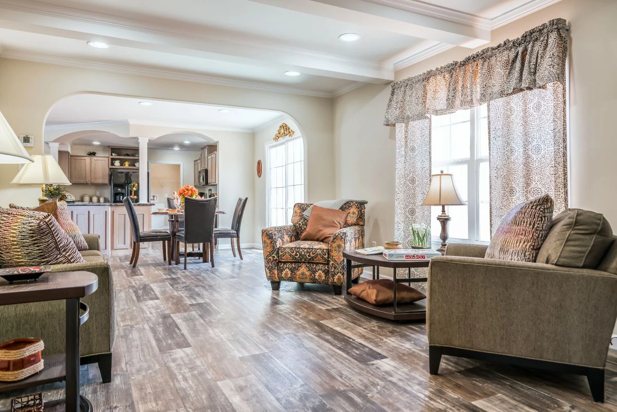 Bright living room with neutral-toned furniture, patterned armchair, wooden floor, coffee table, and large windows, leading to an open kitchen and dining area in the background.