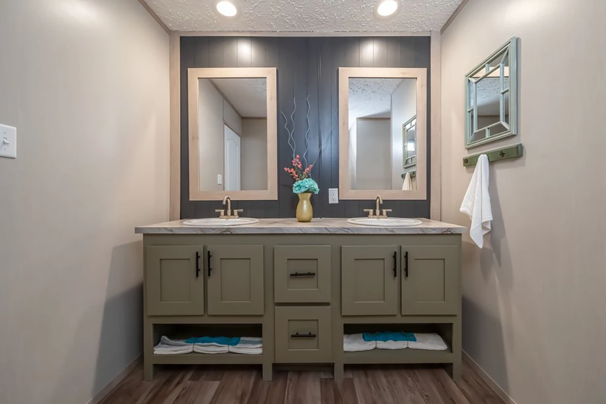 Double bathroom vanity with two sinks, mirrors, a vase with flowers, towel racks, and shelves holding folded towels, set against beige walls and wood flooring.