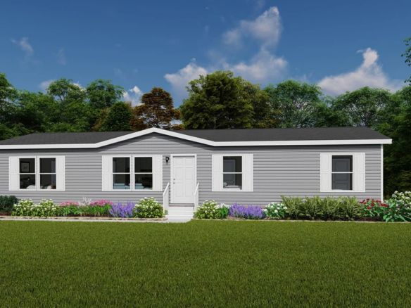 A single-story, gray manufactured home with white trim and a central white door, surrounded by a green lawn, flower beds, and trees in the background under a partly cloudy sky.