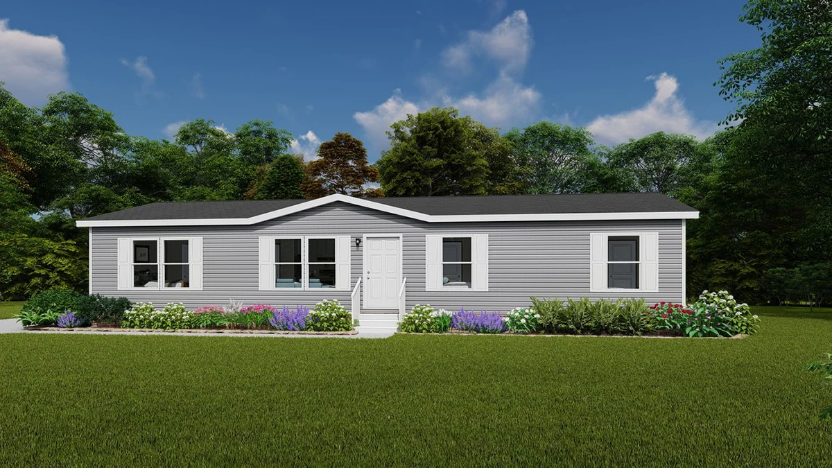 A single-story, gray manufactured home with white trim and a central white door, surrounded by a green lawn, flower beds, and trees in the background under a partly cloudy sky.
