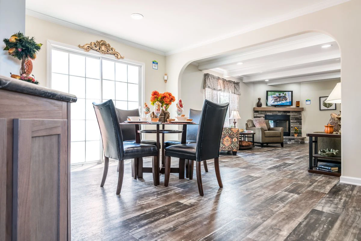 A dining area with a round table and four black chairs is next to a bright living room with sofas, a fireplace, and a TV. Natural light fills the space through large glass doors.