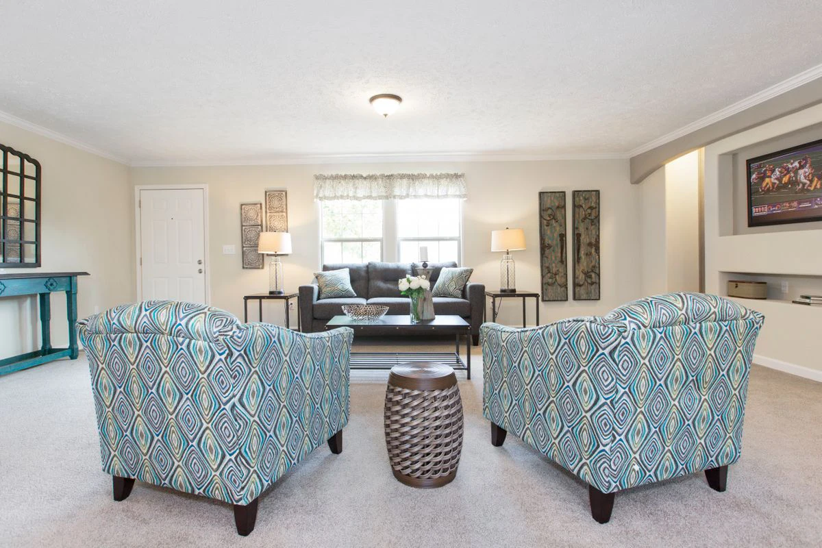 486795ec-393d-443e-ab9d-106c70145ef1 A bright living room with two patterned armchairs, a woven side table, a dark sofa, lamps, and wall art, decorated in neutral and blue tones.