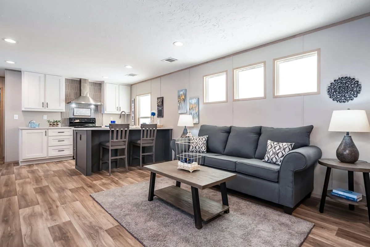 Modern open-concept living room and kitchen with gray sofa, coffee table, bar stools, wood flooring, and natural light from four rectangular windows.