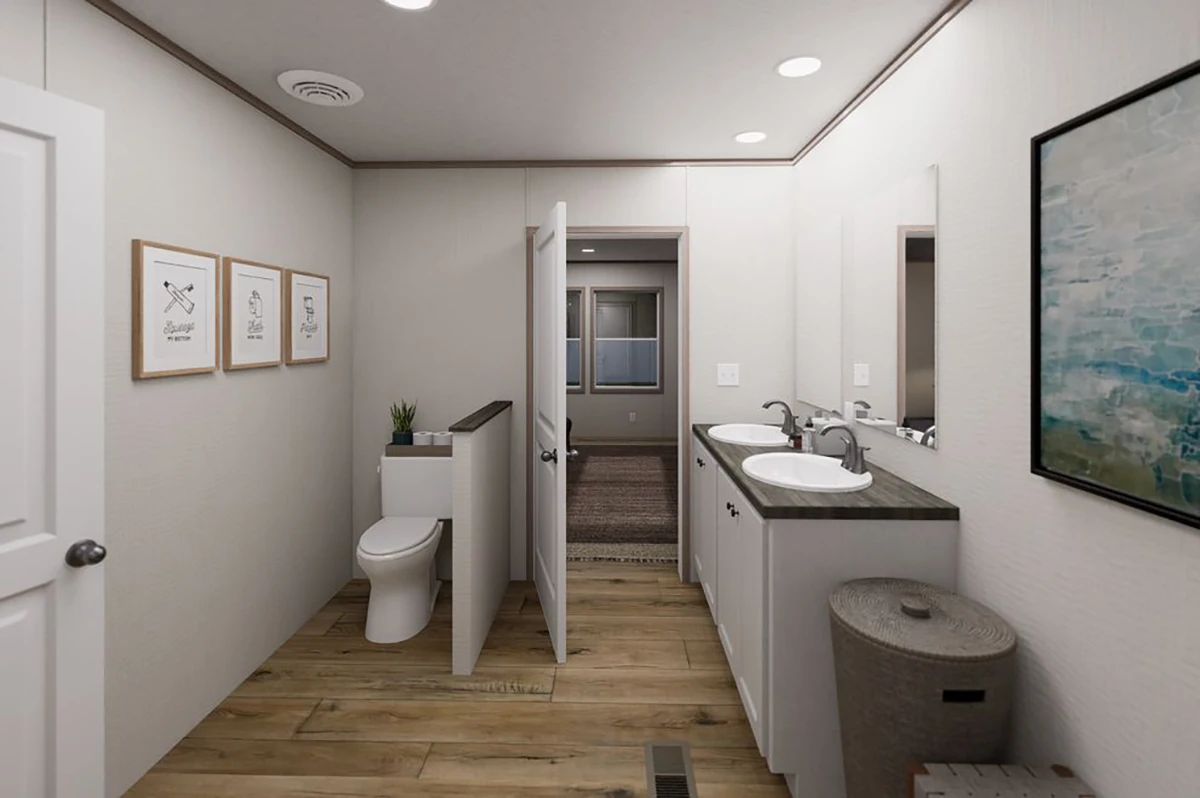 49b88ee7-3aa0-4a26-b633-799903d3ac71 Modern bathroom with double sinks, a toilet beside a half wall, wall art, wood flooring, laundry basket, and recessed lighting. A door is open to reveal an adjoining room.