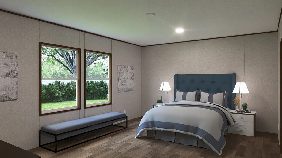 A bedroom with a blue upholstered bed, two nightstands with lamps, a bench under two large windows, and wood flooring. The windows show trees and greenery outside.