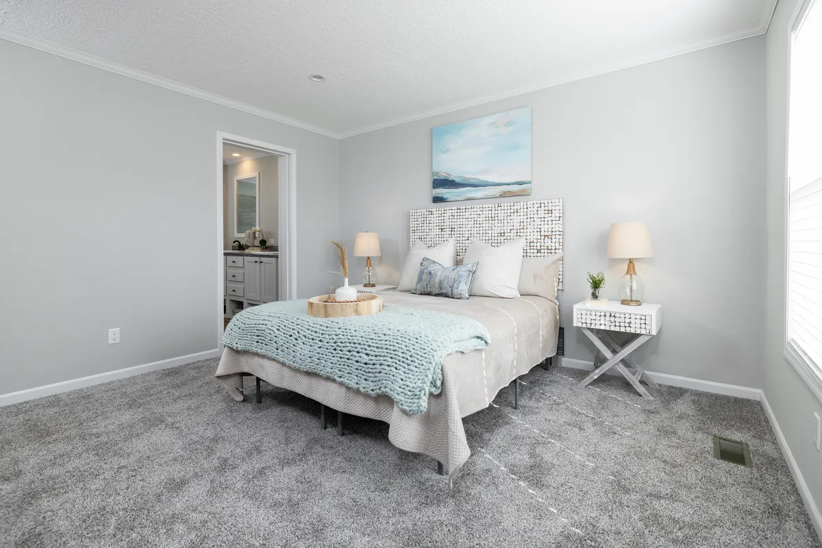 4dd413a1-187a-4800-9ed9-e9d00b3092b7 A neatly made bed with a mosaic headboard, two bedside tables with lamps, and a light blue blanket in a bright, carpeted bedroom with gray walls.