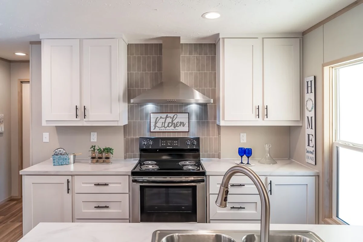 Modern kitchen with white cabinets, stainless steel oven and range hood, tiled backsplash, sink in the foreground, and decorative items on the counter.