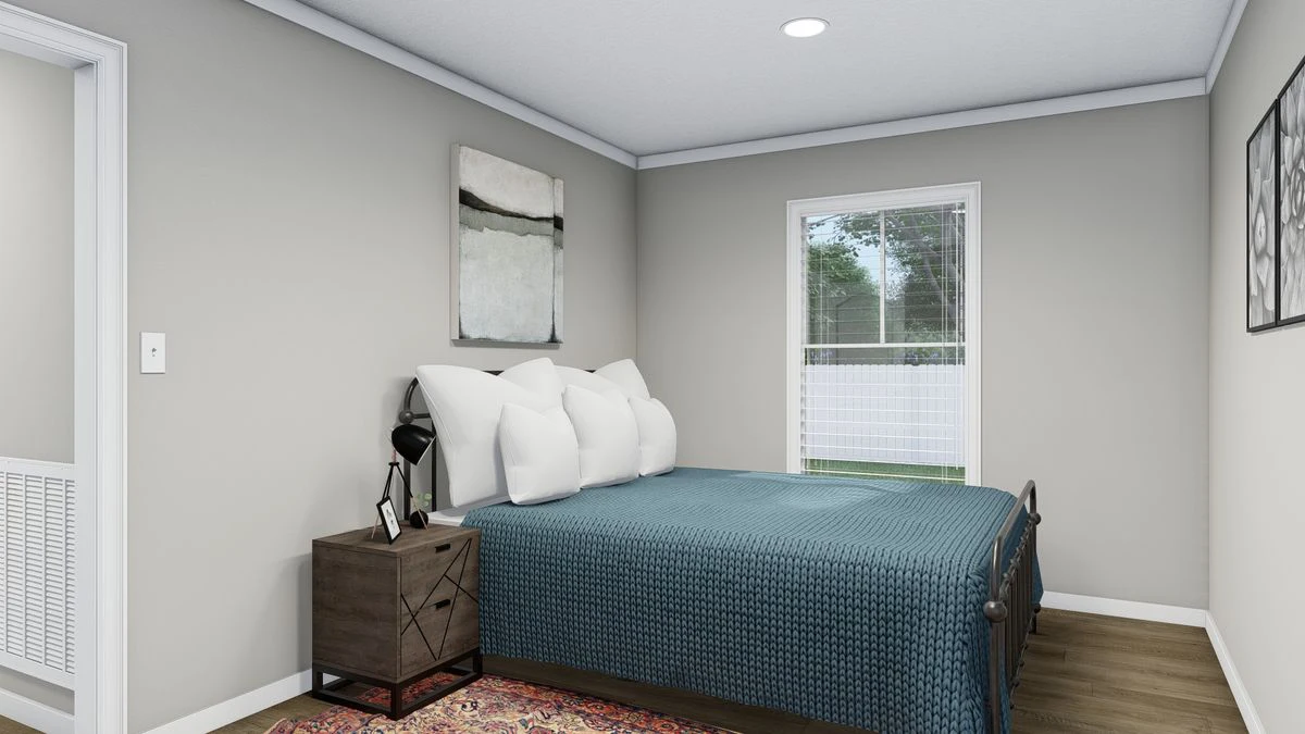 A small bedroom with a bed covered in a blue quilt, four white pillows, a nightstand with a lamp, a window with blinds, and a painting above the bed.