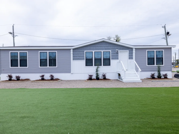 A modern gray and white manufactured home with multiple windows, white front steps, and minimal landscaping, set on artificial turf.