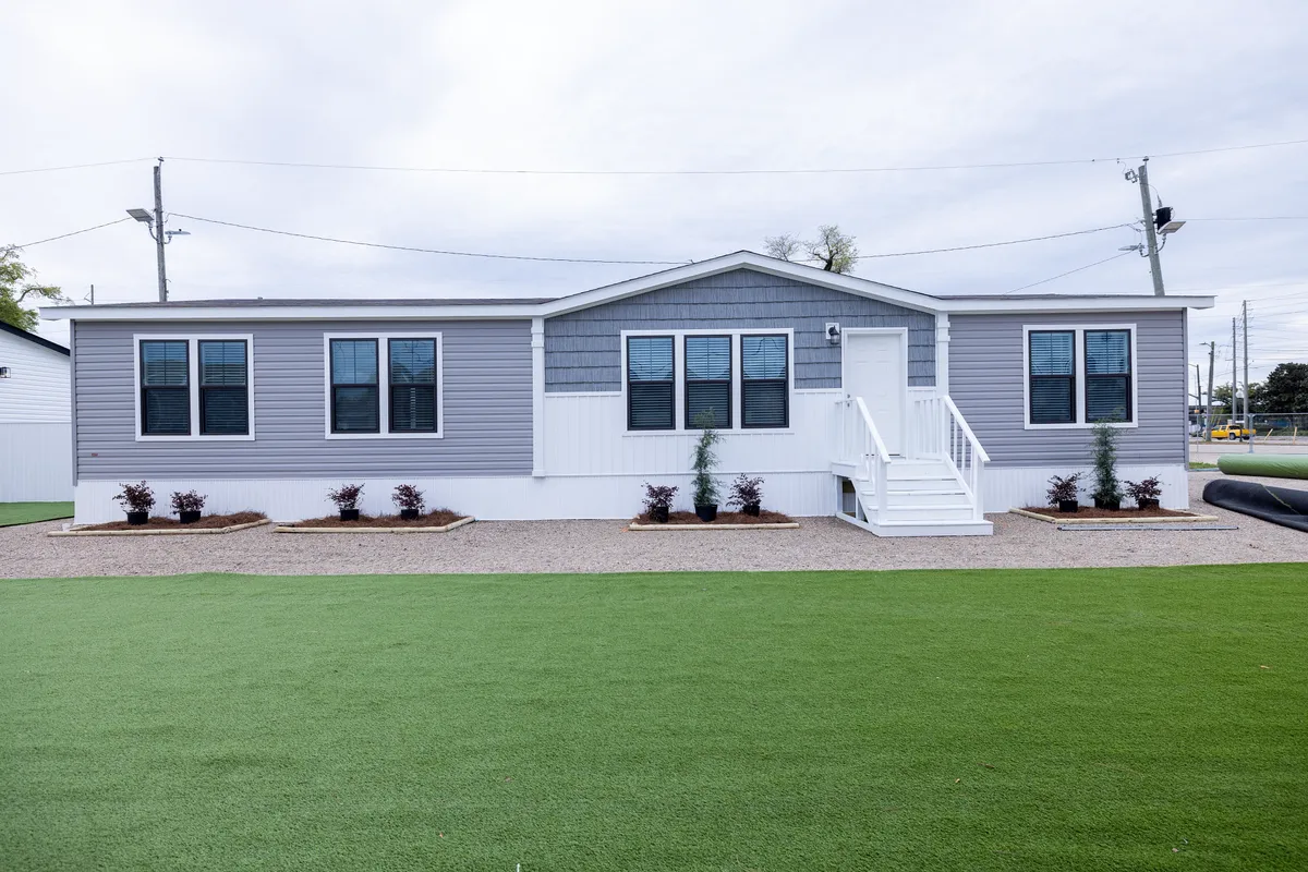 5f89042b-7cae-4595-a73c-24f2693ddd56 A modern gray and white manufactured home with multiple windows, white front steps, and minimal landscaping, set on artificial turf.