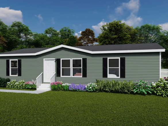 Single-story manufactured home with green siding, black shutters, and white trim, surrounded by shrubs and flowers, set in a grassy yard with a white fence and trees in the background.