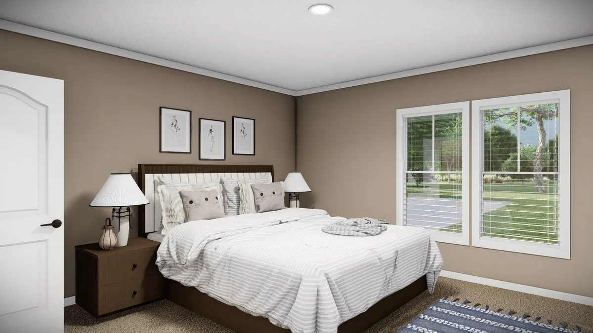 A neatly made bed with white bedding, two brown nightstands with lamps, framed art above the headboard, and two large windows showing a green outdoor view.