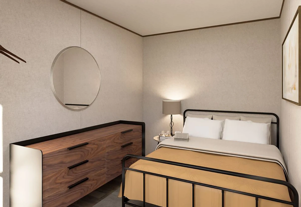 A small bedroom with a metal bed frame, tan bedding, a wooden dresser with a round mirror, a lamp, and a framed picture on the wall.