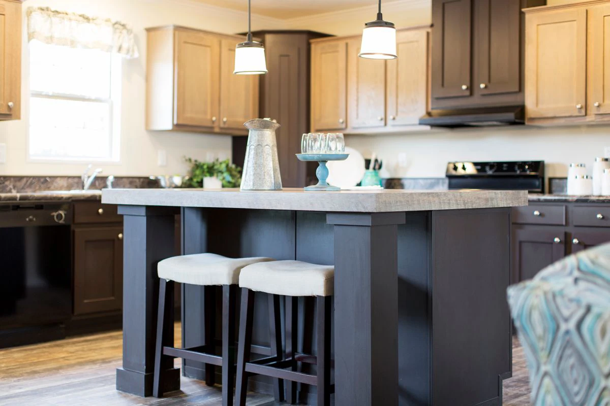 6d6f33ea-a943-4909-a738-f617ef1da688 Modern kitchen with a central island, two upholstered stools, a metal pitcher, glasses on a tray, and light brown cabinetry under pendant lights.
