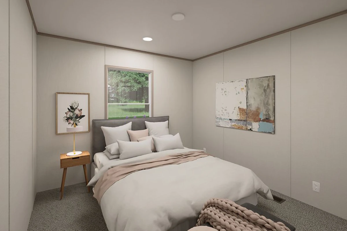 7170c1be-183c-4791-b225-77942c84e5e6 A small bedroom with a bed, side table, framed artwork, and a window showing greenery outside. The walls are light-colored and the floor is carpeted.
