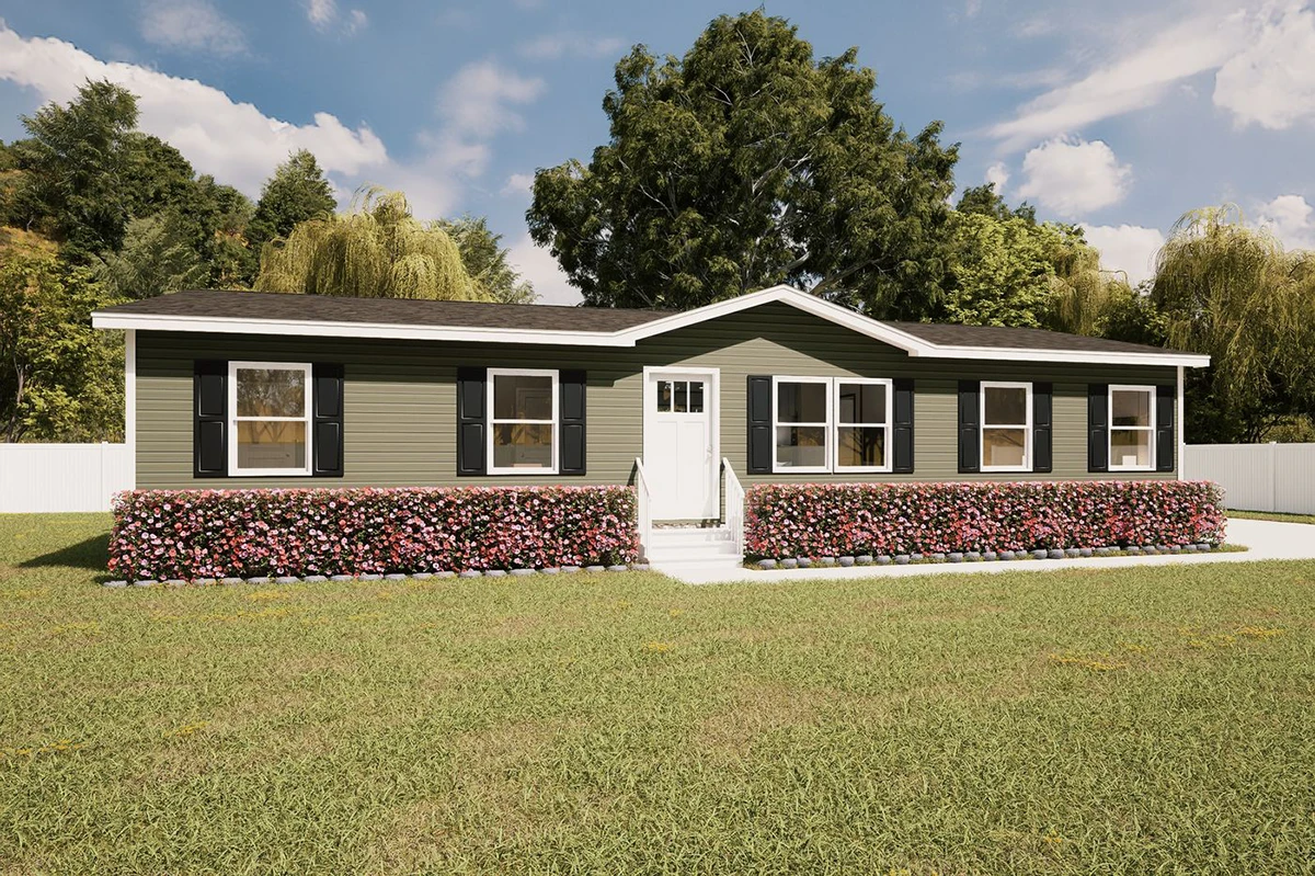 72cd4ebf-9b19-4f9a-863f-f36029d3beea A single-story green manufactured home with black shutters, white door, front steps, a hedge with pink flowers, and a lawn, set against trees and a blue sky.