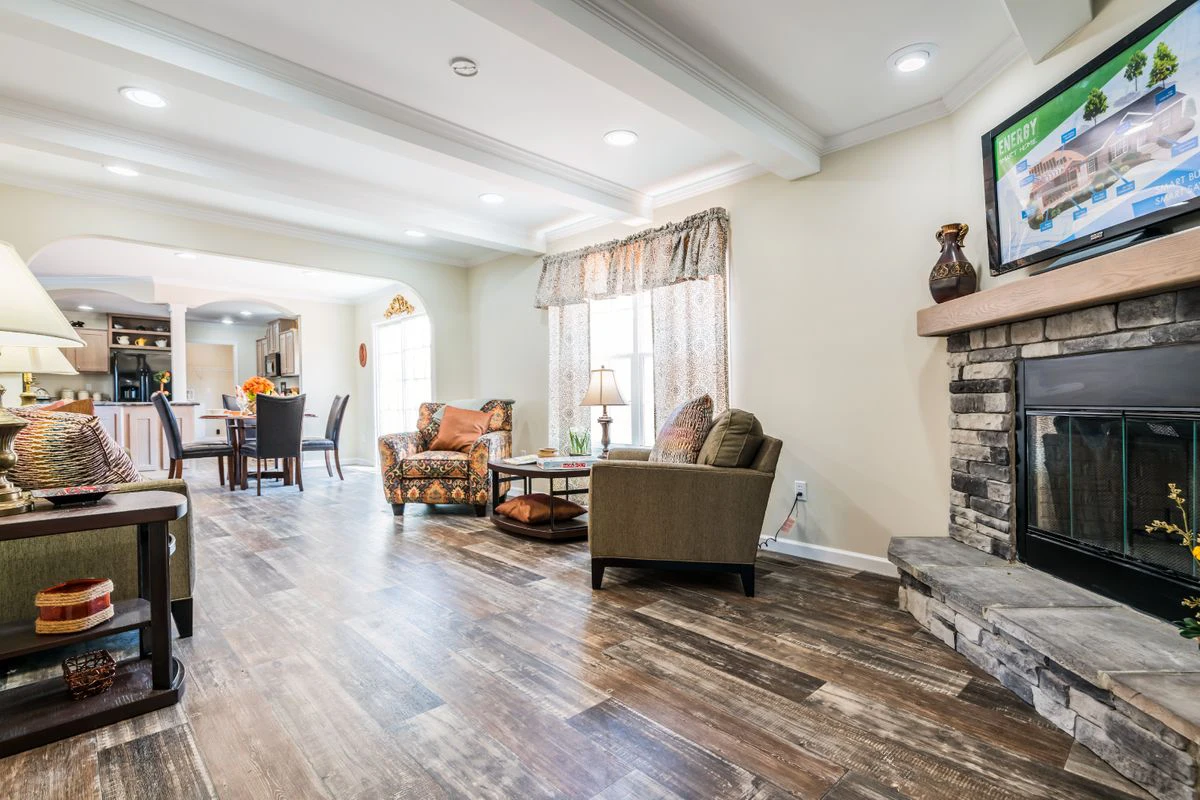 Bright living room with wood-look flooring, stone fireplace, armchairs, and TV above mantle; dining area and kitchen visible in the background.
