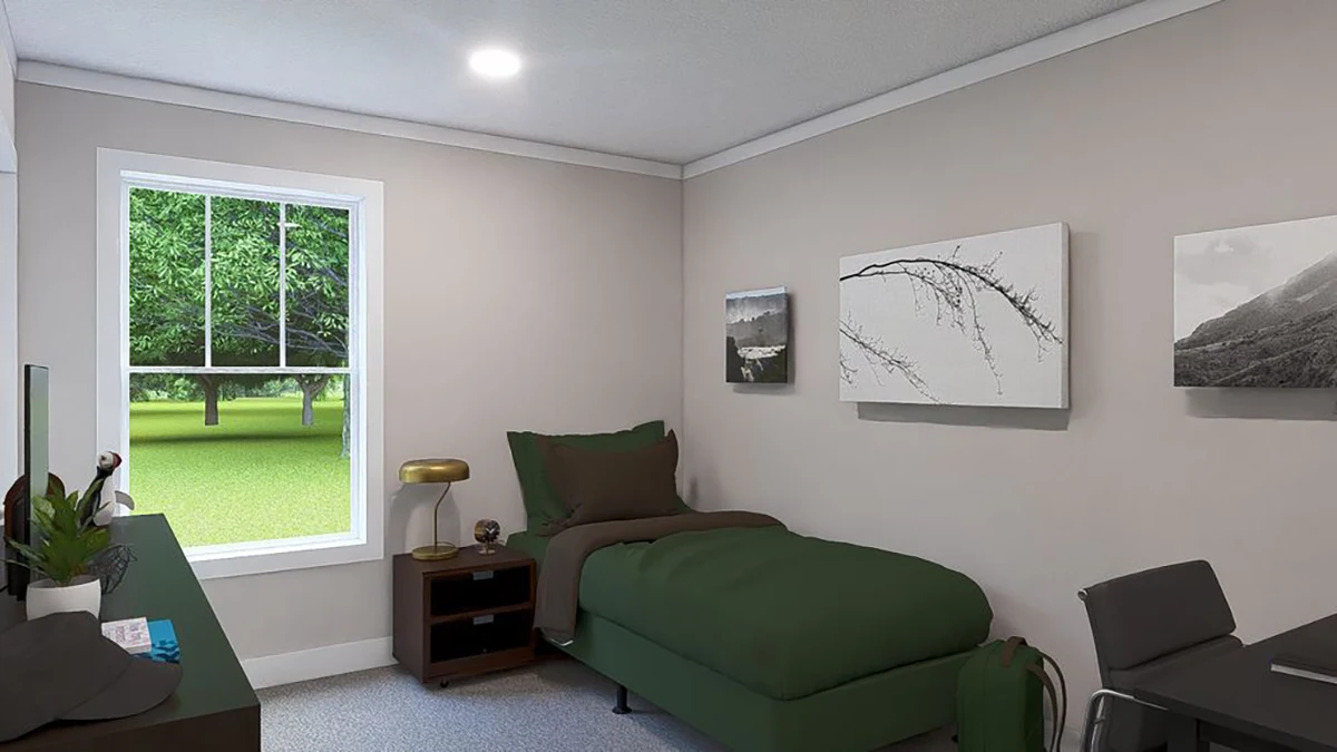 A small bedroom with a single bed, green bedding, a nightstand with a lamp, a desk with a chair, wall art, and a window showing a grassy outdoor area with trees.