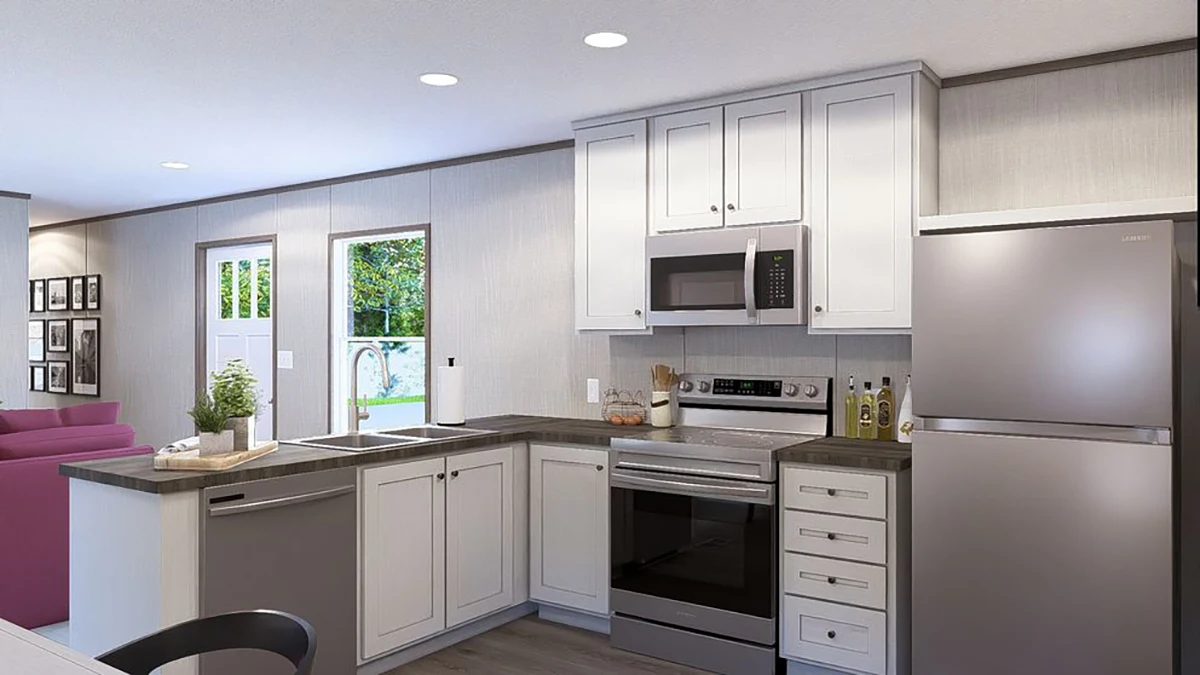 Modern kitchen with white cabinets, stainless steel appliances, gray countertops, and a window above the sink; adjacent living area visible in the background.
