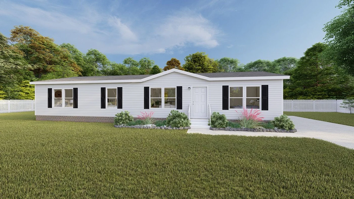 7df4d3ee-a587-490d-adcb-f70787d5ef65 Single-story white manufactured home with black shutters, front lawn, small garden beds, and a driveway, set against a background of trees under a partly cloudy sky.