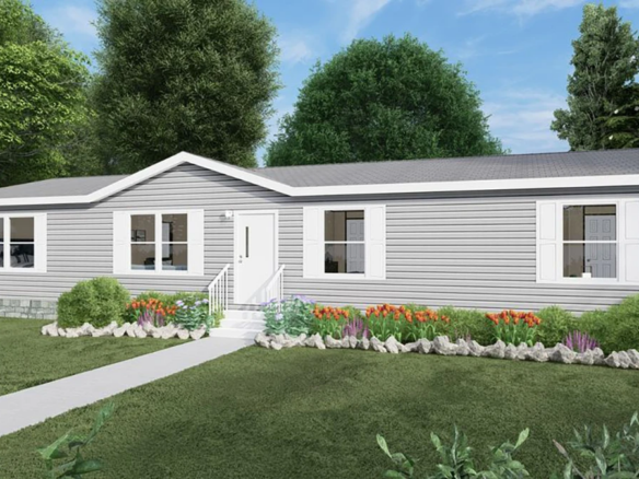 Single-story gray manufactured home with white trim, front steps, and a landscaped garden with flowers and shrubs, surrounded by trees and grass under a blue sky.