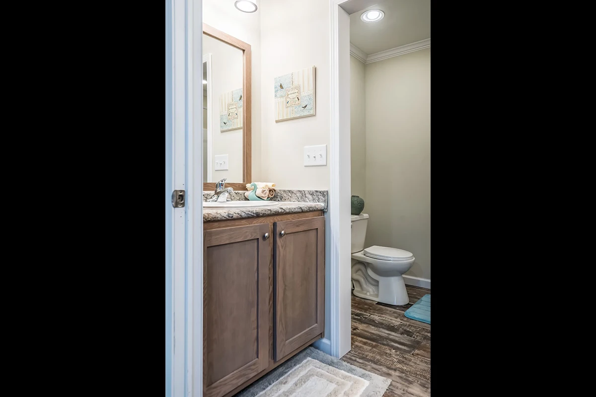 A bathroom with a wooden vanity, granite countertop, mirror, wall art, toilet, and wood-look flooring. Blue and white rugs are on the floor.