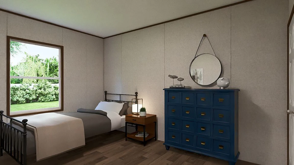 A bedroom with a single metal bed, a wooden nightstand, a blue dresser with a round mirror above it, and a window showing greenery outside.