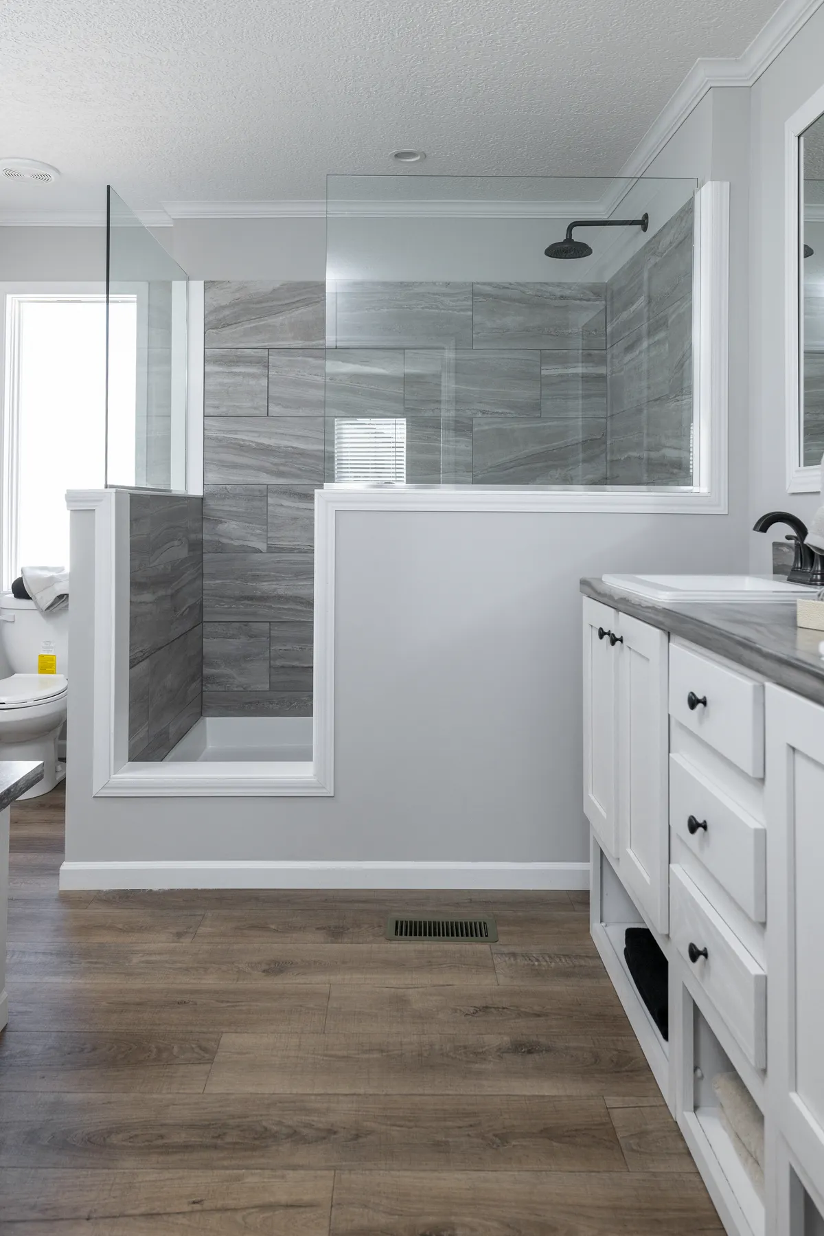 8f33cd08-9550-45d6-ba89-1e67a62dc1e3 Modern bathroom with a glass-enclosed shower featuring gray tile, white vanity with drawers, and wood-look flooring.
