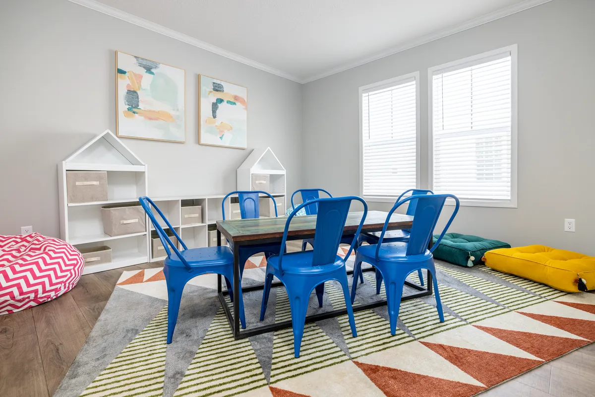 917227ae-e1a6-4525-a002-628741232bb9 A brightly lit playroom with a table, six blue chairs, colorful geometric rug, bean bag, cushions, two house-shaped shelves, and abstract art on light gray walls.
