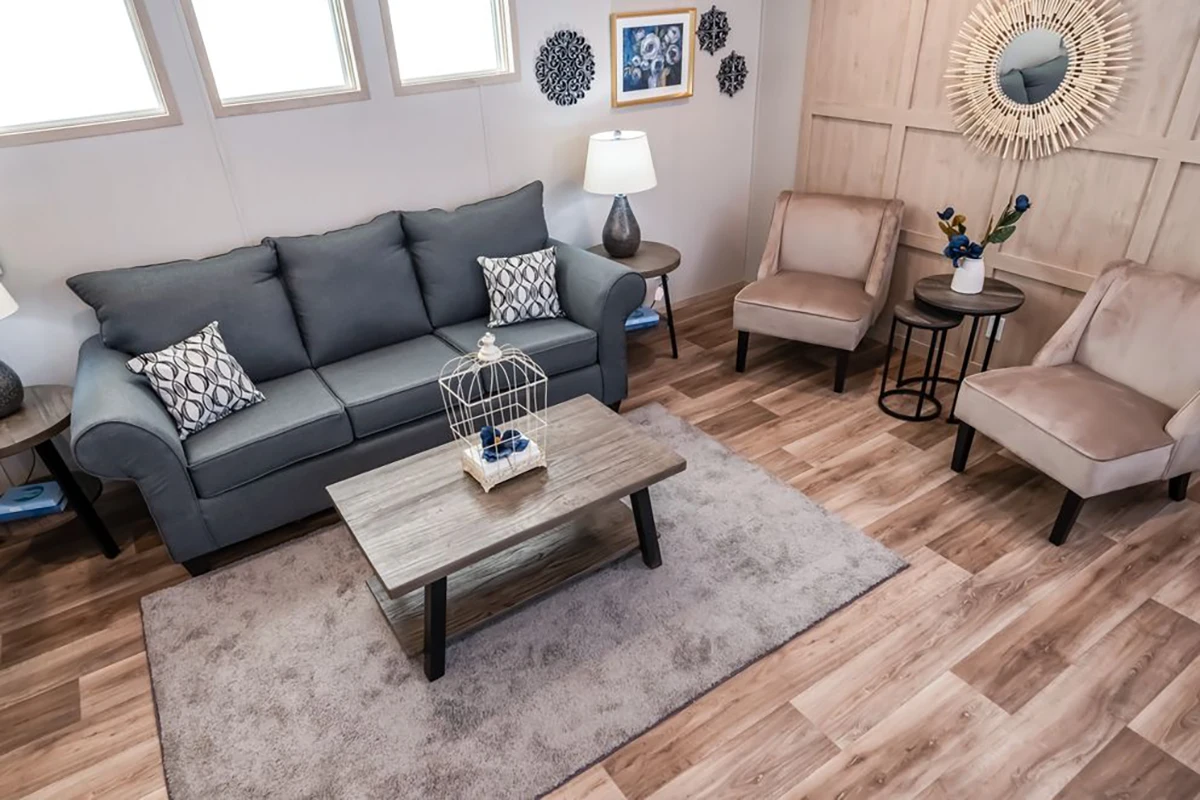 A modern living room with a gray sofa, two beige chairs, a wooden coffee table, decorative wall art, and a light-colored wood floor. Three windows provide natural light.