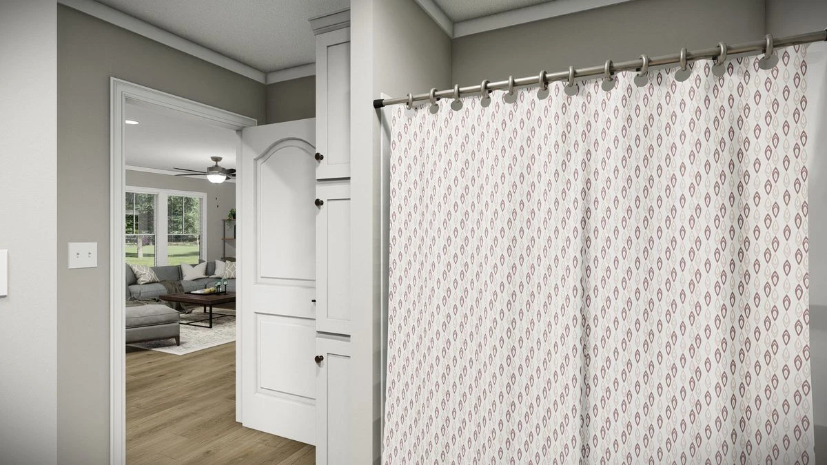 94e588ff-5b9f-47c8-9b09-803c30348c7f A bathroom with a patterned shower curtain, partial view of white cabinets, and an open door leading to a living room with a sofa and windows.
